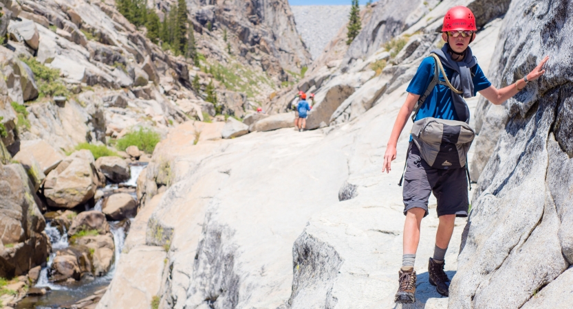 A student wearing a helmet braces themself on a rock wall while making their way across narrow rocky terrain. There is a stream below them.